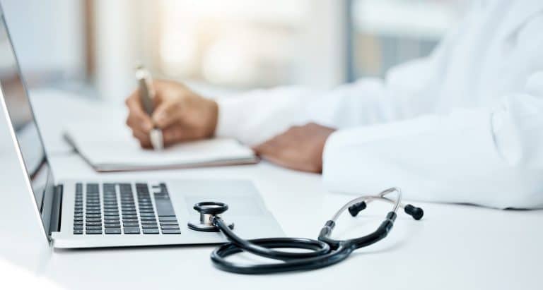 Doctor writing on notepad in front of open laptop